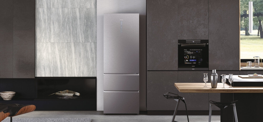 W kuchni znajduje się lodówka w kolorze platynowy inox z dwoma dolnymi szufladami. Obok lodówki widać zabudowany piekarnik z wyświetlaczem.