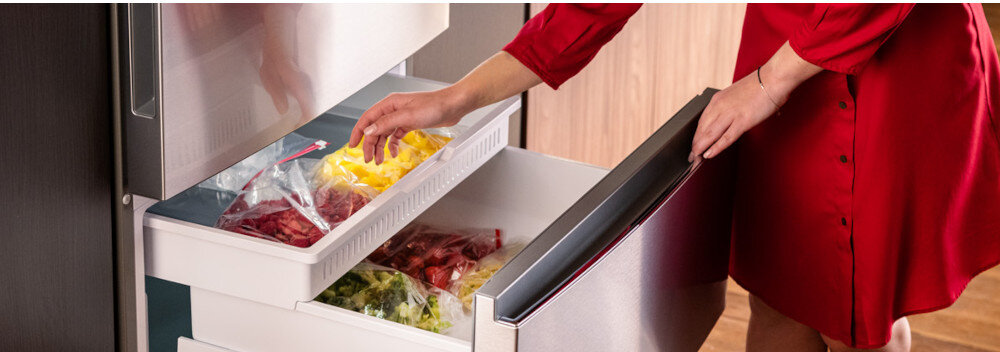 Osoba w czerwonym ubraniu otwiera dolną szufladę lodówki, w której znajdują się zapakowane w folię kawałki owoców i warzyw. Lodówka ma srebrny kolor i nowoczesny design.