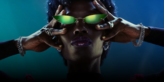 Osoba nosi zielone okulary przeciwsłoneczne o nietypowym kształcie, trzymając je obiema rękami. Tło jest ciemne z niebiesko-zielonym oświetleniem.