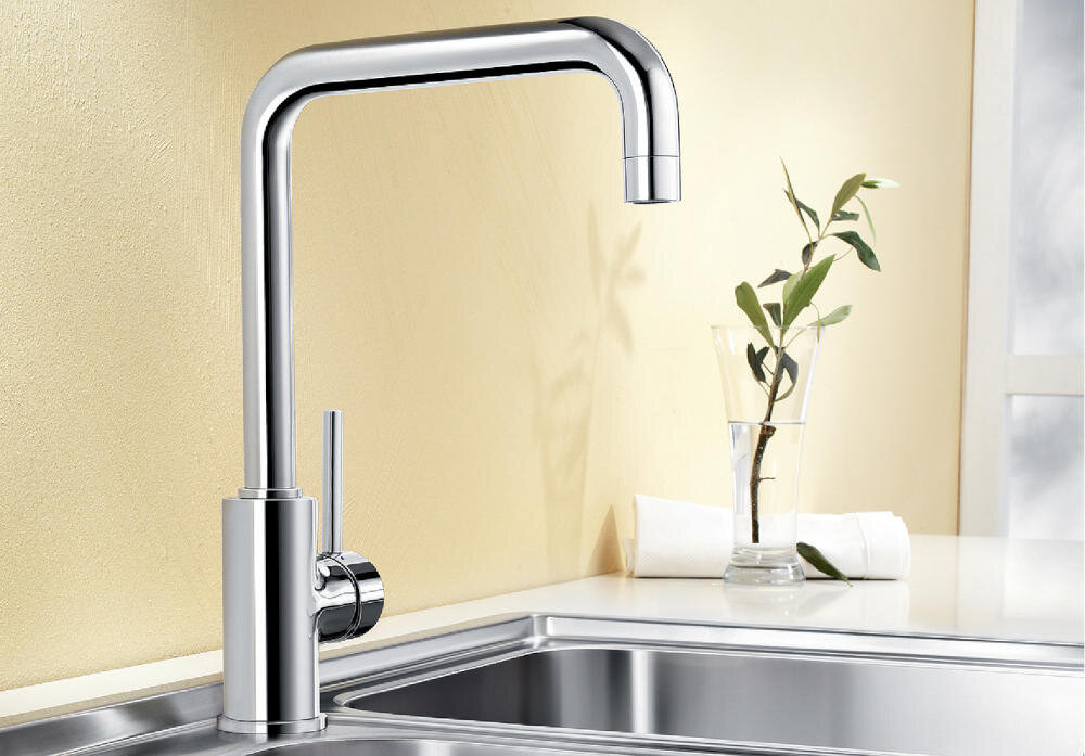 Chromowana bateria kuchenna o prostokątnym kształcie z dźwignią po boku, zamontowana nad zlewem. W tle widać jasną ścianę i szklankę z gałązką rośliny.