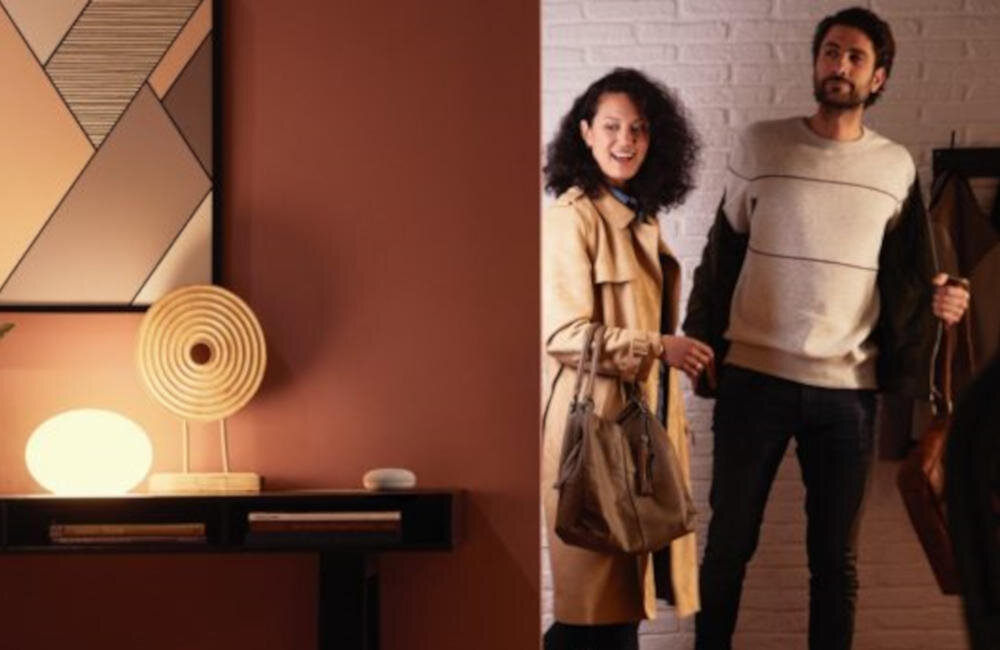 Wnętrze pokoju z lampą na stoliku, obok której znajduje się dekoracyjny przedmiot. Dwie osoby stoją przy ścianie z cegieł, jedna z nich trzyma torbę.