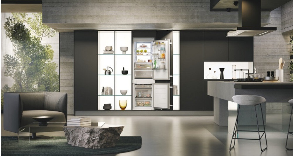 Nowoczesna kuchnia z otwartą lodówką w zabudowie, w której widać różne produkty spożywcze. Wnętrze kuchni jest eleganckie, z ciemnymi szafkami i wyspą kuchenną, a na półkach znajdują się dekoracyjne przedmioty.