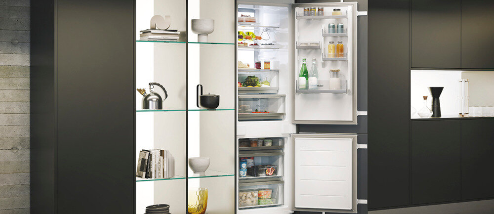 Otwarta lodówka z widocznymi półkami wypełnionymi różnymi produktami spożywczymi, takimi jak butelki, słoiki i warzywa. Obok lodówki znajduje się regał z dekoracyjnymi przedmiotami, takimi jak książki i naczynia.
