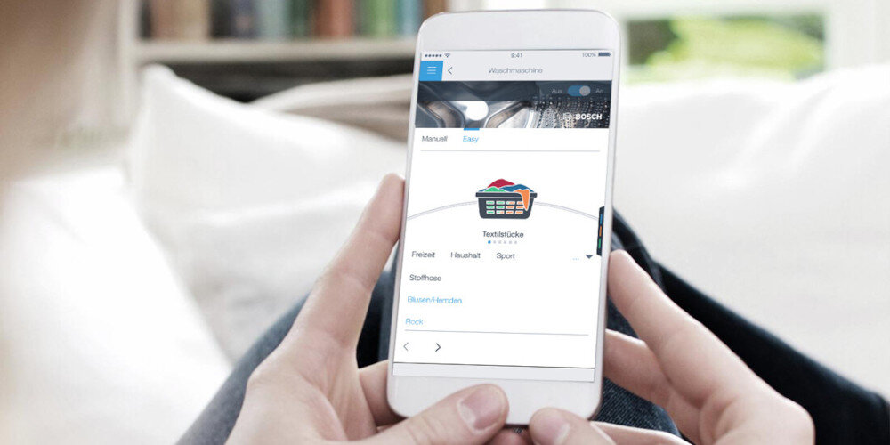 Osoba trzyma smartfon, na którym wyświetlana jest aplikacja z logo BOSCH i opcjami wyboru trybu prania. Widoczny jest koszyk z kolorowym praniem oraz tekst: \'Textilpflege\', \'Freizeit\', \'Haushalt\', \'Sport\'.