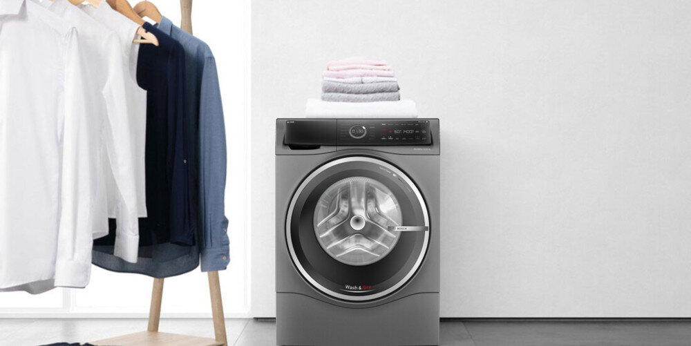 Na obrazie znajduje się pralko-suszarka w kolorze szarym z widocznym okrągłym bębnem i panelem sterowania. Na urządzeniu leży stos złożonych ręczników, a obok widać stojak z powieszonymi koszulami.