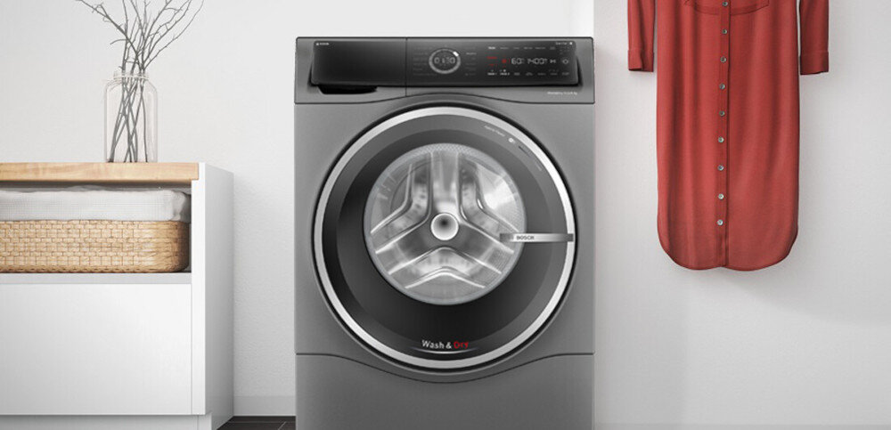 Pralko-suszarka w kolorze szarym z widocznym bębnem i panelem sterowania. Obok znajduje się kosz z ręcznikami i wisząca czerwona koszula.