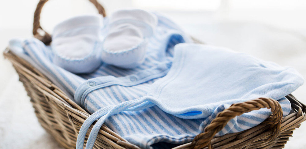 Koszyk wiklinowy zawiera niebieskie ubranka dziecięce i białe buciki. Ubranka mają wzór w paski.