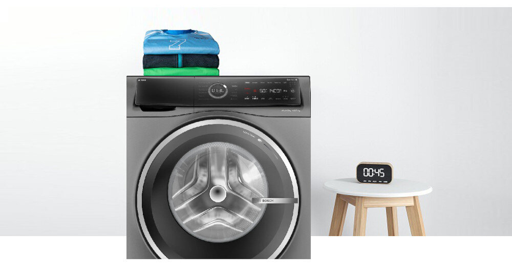 Pralko-suszarka w kolorze srebrnym z widocznym logo BOSCH na drzwiach. Obok na stoliku stoi zegar elektroniczny z wyświetlaczem pokazującym \'00:45\'.