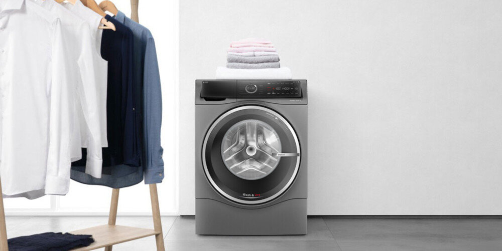 Na obrazie znajduje się pralko-suszarka w kolorze srebrnym z widocznym panelem sterowania i okrągłym bębnem. Obok stoi wieszak z powieszonymi koszulami, a na urządzeniu leży stos złożonych ręczników.