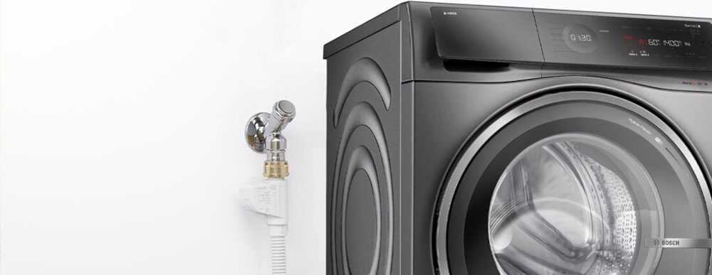 Na obrazie widoczna jest pralko-suszarka w kolorze ciemnym z okrągłym bębnem i panelem sterowania. Obok urządzenia znajduje się przyłącze wodne z wężem.