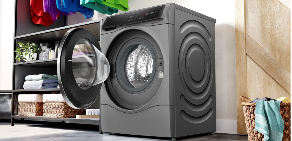 Na obrazie znajduje się szara pralko-suszarka z otwartymi drzwiami. W tle widoczne są półki z kolorowymi ubraniami i koszami.