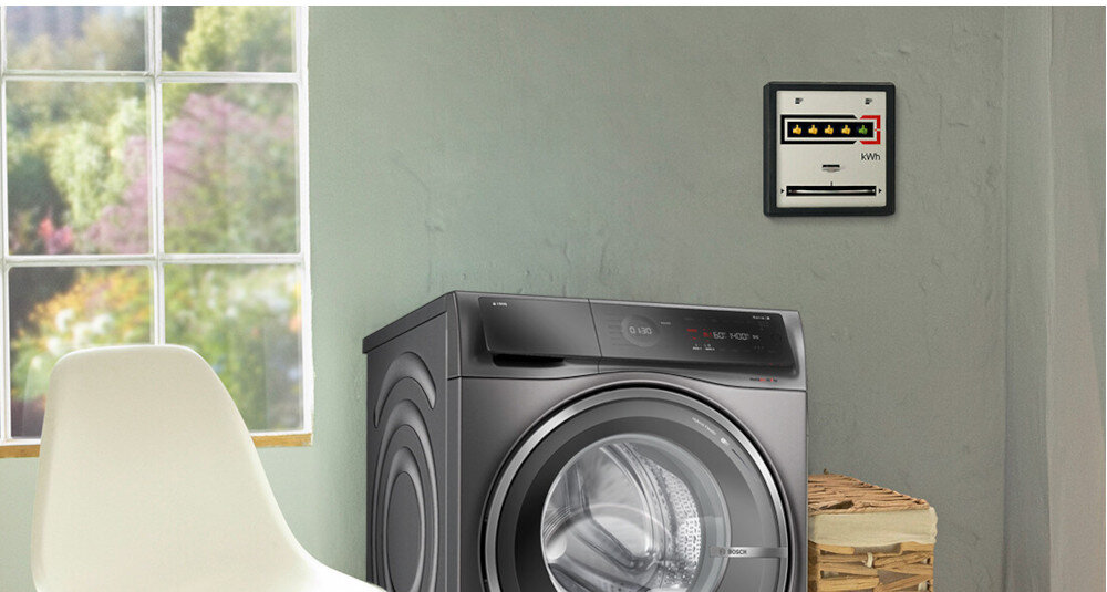 Na obrazie znajduje się pralko-suszarka w kolorze ciemnoszarym, ustawiona obok wiklinowego kosza. W tle widać okno z widokiem na zieleń oraz licznik energii na ścianie z oznaczeniem \'kWh\'.