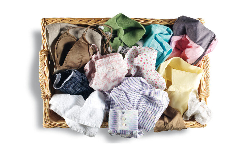 Kosz wiklinowy wypełniony różnokolorowymi ubraniami, w tym koszulami, bluzkami i bielizną. Ubrania są złożone i ułożone w sposób przypadkowy.
