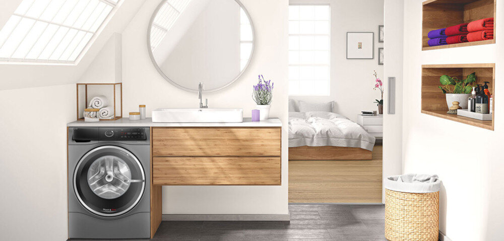 W jasnej łazience znajduje się pralko-suszarka z widocznym bębnem, obok drewniana szafka z umywalką i okrągłym lustrem. W tle widać sypialnię z łóżkiem oraz półki z ręcznikami i kosmetykami.