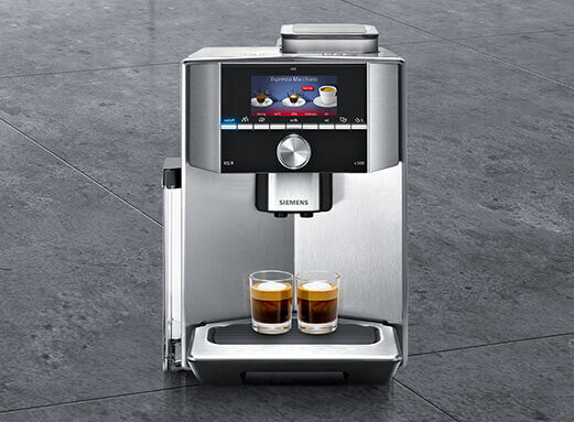 Srebrny ekspres do kawy z wyświetlaczem pokazującym różne rodzaje kawy. Na tacy znajdują się dwa szklane kubki z kawą, a poniżej widoczne jest logo \'SIEMENS\'.