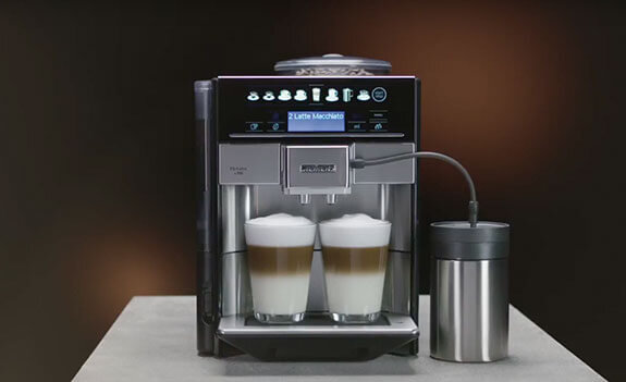 Ekspres do kawy z wyświetlaczem pokazującym napis \'2 Latte Macchiato\', z dwoma filiżankami wypełnionymi kawą z mleczną pianką. Obok ekspresu znajduje się metalowy pojemnik z rurką.