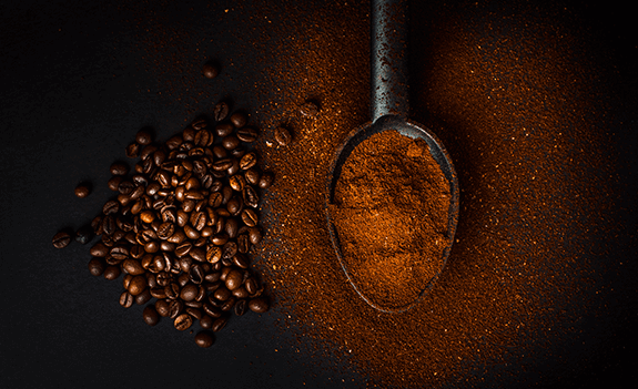 Na ciemnym tle widoczne są ziarna kawy oraz metalowa łyżka wypełniona mieloną kawą.