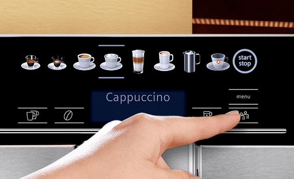 Panel sterowania ekspresu do kawy z ikonami różnych napojów, przyciskiem \'start stop\' oraz wyświetlaczem z napisem \'Cappuccino\'. Dłoń wskazuje na przycisk na panelu.