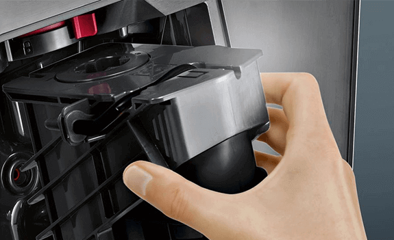 Osoba trzyma czarny element wewnętrzny ekspresu do kawy, widoczny jest fragment mechanizmu urządzenia.