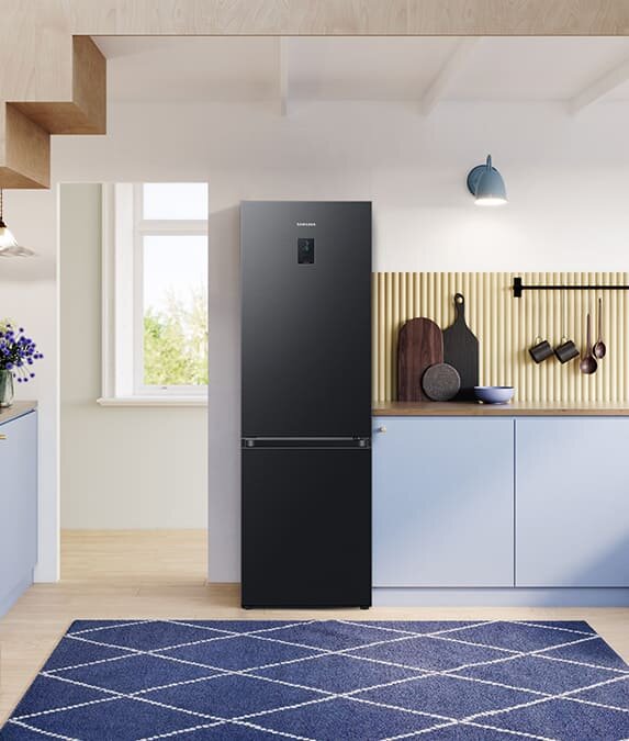 Ponadczasowy wygląd łączy estetykę z wygodą użytkowania Czarna lodówka z logo Samsung stoi w nowoczesnej kuchni z jasnymi szafkami i drewnianym blatem. Na blacie znajdują się deski do krojenia i miska, a na ścianie wisi lampa i kilka przyborów kuchennych.