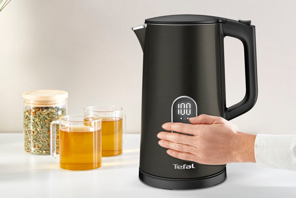 Czarny czajnik elektryczny z wyświetlaczem pokazującym temperaturę 100°C i logo Tefal na dole. Obok znajdują się dwie szklanki z napojem oraz słoik z suszonymi ziołami.