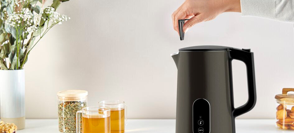 Czarny czajnik elektryczny z regulacją temperatury stoi na blacie, obok dwóch szklanek z herbatą i słoika z suszonymi ziołami. Ręka trzymająca mały przedmiot znajduje się nad czajnikiem.