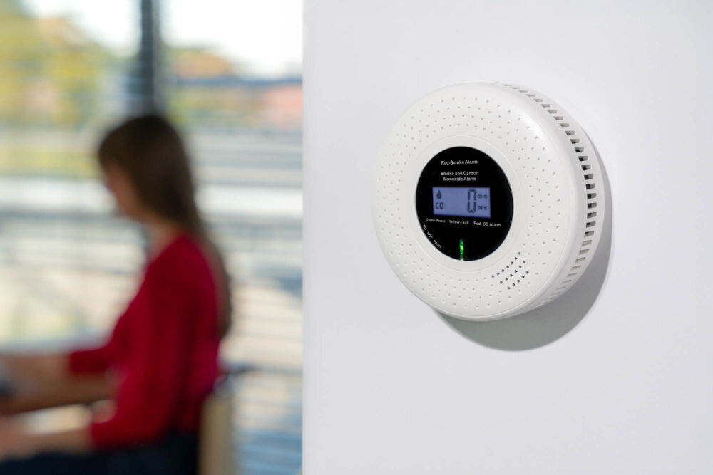 CZUJNIK DYMU I TLENKU WĘGLA VÖGEL VCA200 Okrągły, biały czujnik dymu i tlenku węgla (czadu) VÖGEL z cyfrowym wyświetlaczem LCD, zamontowany na jasnej ścianie. Wyświetlacz pokazuje 0 ppm dla stężenia CO. W tle, po lewej stronie, rozmyta postać kobiety w czerwonej koszulce siedzącej przy biurku.