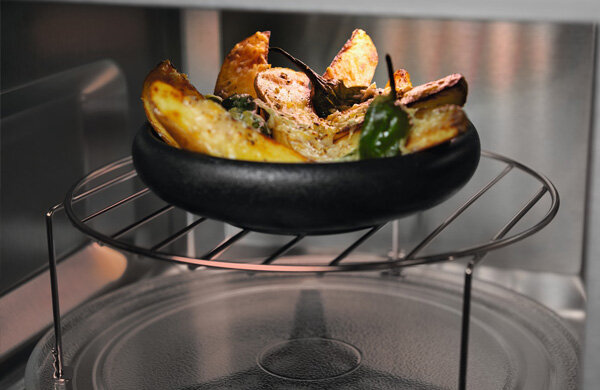 Wnętrze kuchenki mikrofalowej z czarną misą na metalowej kratce, w której znajdują się pieczone ziemniaki i zielone papryczki.