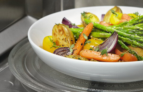 Na obrazie widoczny jest talerz z różnorodnymi warzywami, takimi jak szparagi, marchewki, ziemniaki i cebula, umieszczony wewnątrz kuchenki mikrofalowej. Talerz stoi na szklanym talerzu obrotowym z napisem \'FOR MICROWAVE OVEN ONLY\'.