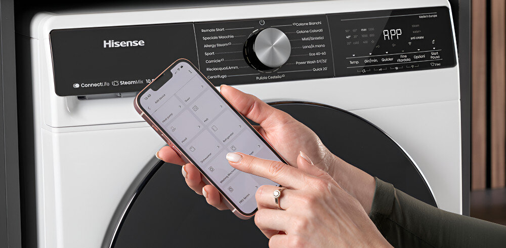 Osoba trzyma smartfon z aplikacją do sterowania suszarką, na której panelu widoczne jest logo Hisense oraz różne opcje programów. Suszarka ma nowoczesny design z dużym pokrętłem i wyświetlaczem cyfrowym.