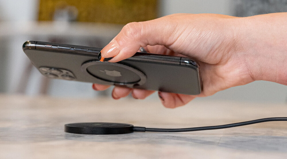 Osoba trzyma smartfon nad czarną ładowarką indukcyjną umieszczoną na stole. Ładowarka jest podłączona kablem do źródła zasilania.