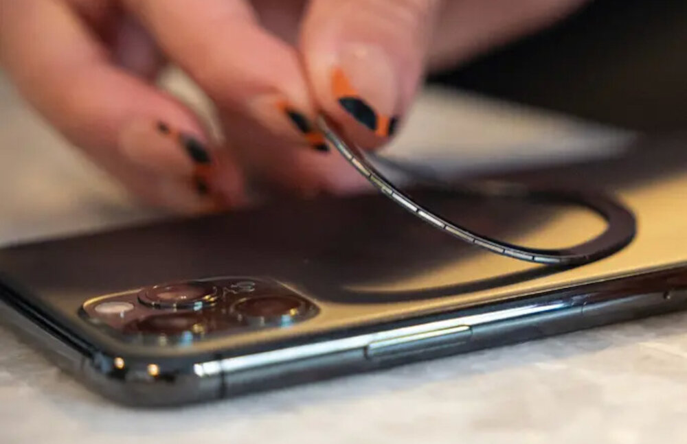 Osoba trzyma cienki, okrągły element nad smartfonem z trzema obiektywami aparatu. Element wydaje się być magnetycznym pierścieniem lub nakładką.