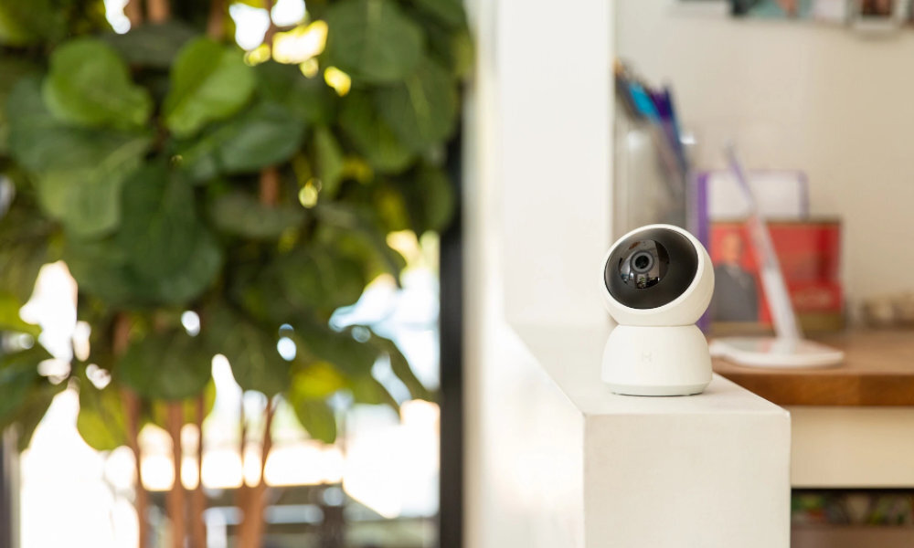 Na białej powierzchni stoi biała, obrotowa kamera wewnętrzna. W tle widoczna jest zielona roślina i rozmyte elementy wnętrza.