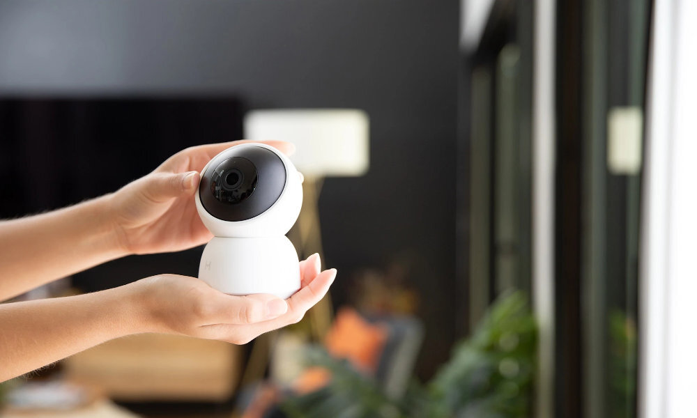 Osoba trzyma białą, obrotową kamerę wewnętrzną z czarnym obiektywem. W tle widoczne są rozmyte elementy wnętrza, takie jak rośliny i lampa.