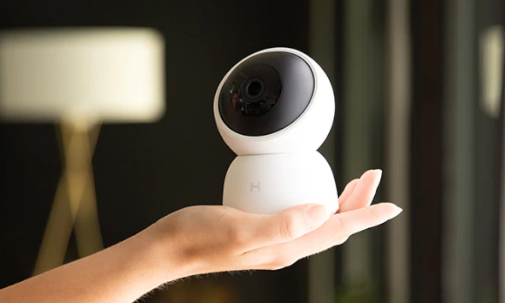 Osoba trzyma w dłoni białą, obrotową kamerę z czarnym obiektywem. W tle widoczna jest rozmyta lampa stojąca.