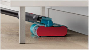 Odkurzacz z turkusowo-czerwoną głowicą czyszczącą znajduje się pod meblem na drewnianej podłodze. Widoczna jest rura odkurzacza oraz część głowicy z przezroczystym elementem.