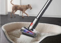 Odkurzacz z metaliczną rurą i głowicą czyszczącą znajduje się na powierzchni legowiska dla zwierząt. W tle widoczny jest pies na ciemnej podłodze.