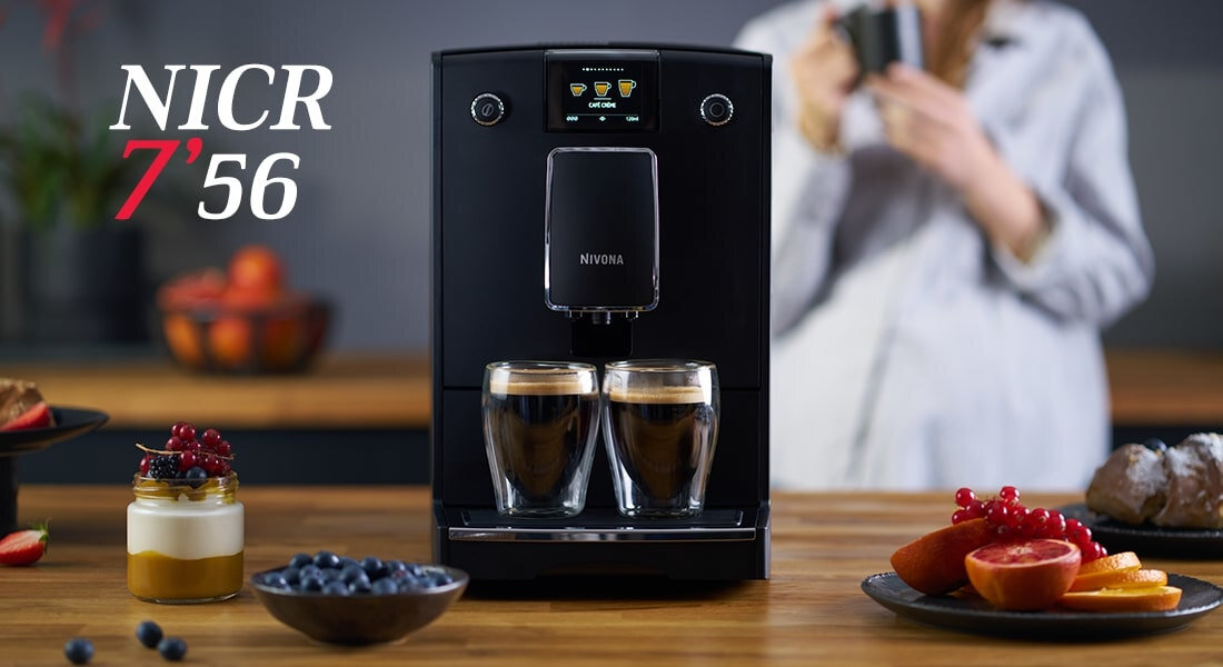 Na blacie stoi czarny ekspres do kawy z logo NIVONA, przed nim dwie szklanki z kawą. Obok znajdują się owoce i deser, a w tle osoba trzyma filiżankę.