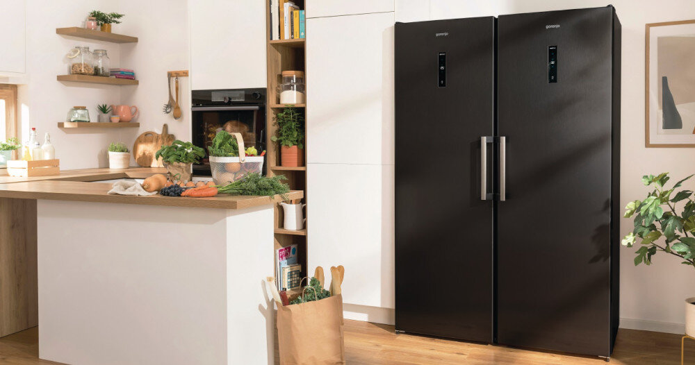 Kuchnia z czarną zamrażarką i lodówką marki Gorenje, obok drewniany blat z warzywami i torba z zakupami. Na półkach znajdują się rośliny i naczynia.