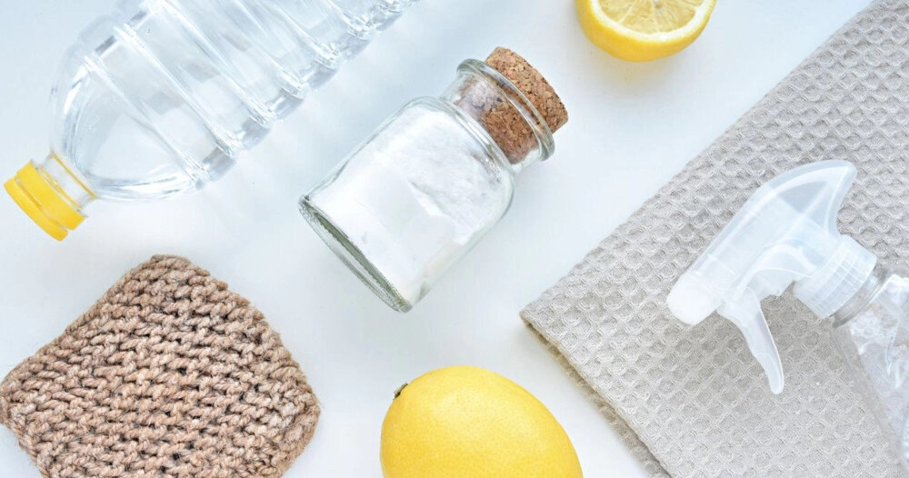 Na białym tle znajdują się plastikowa butelka z przezroczystym płynem, szklany słoik z białym proszkiem, cytryna, połówka cytryny, brązowa szorstka ściereczka i plastikowy spryskiwacz. Obok leży jasnoszara teksturowana ściereczka.