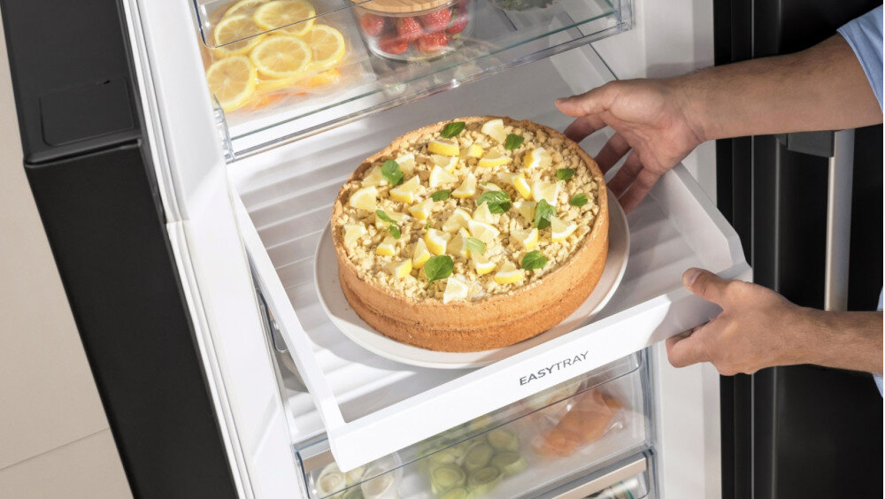 Osoba wkłada ciasto na półkę oznaczoną napisem \'EASYTRAY\' w zamrażarce. W tle widoczne są inne produkty spożywcze, takie jak plasterki cytryny i pomidory.