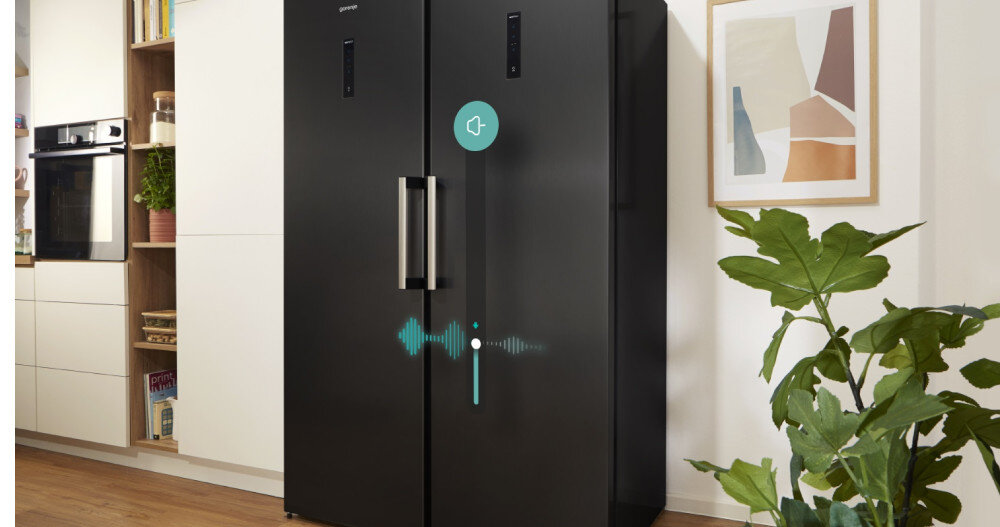 Czarna zamrażarka z dwoma pionowymi uchwytami stoi w nowoczesnej kuchni. Na drzwiach widoczne są cyfrowe wskaźniki i ikona głośnika z falą dźwiękową.