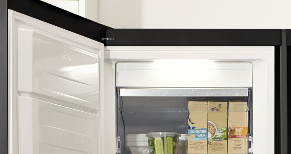 Otwarta zamrażarka z widocznymi produktami spożywczymi w środku, w tym kartonami i pojemnikiem z zielonymi warzywami. Na górnej krawędzi drzwi widoczne jest logo \'ADAPTTECH\'.