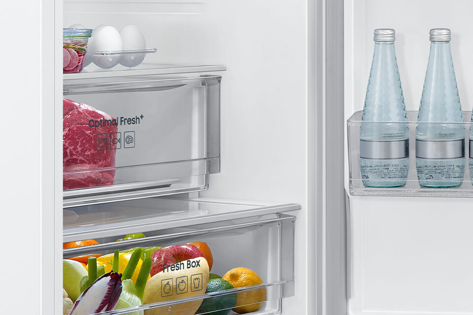 Wnętrze lodówki z widocznymi półkami i szufladami. Na półkach znajdują się jajka, mięso, warzywa, owoce oraz butelki z wodą; widoczny jest napis \'Optimal Fresh+\' i \'Fresh Box\'.