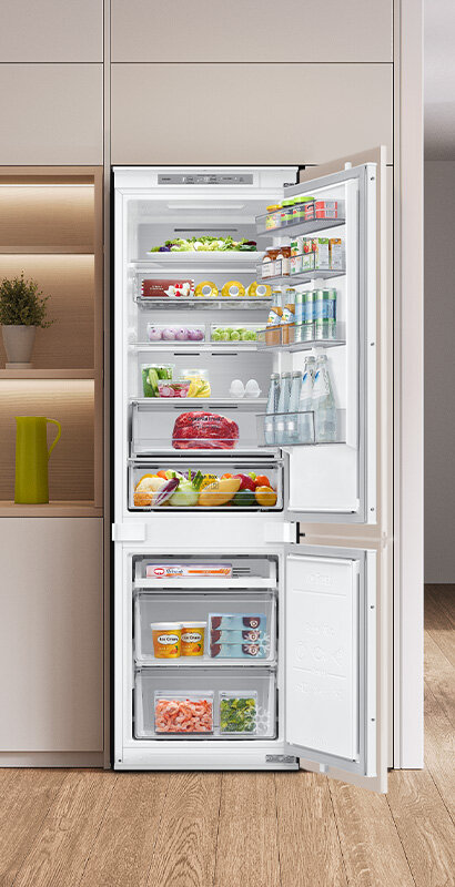 Otwarta lodówka z widocznymi półkami wypełnionymi różnorodnymi produktami spożywczymi, takimi jak warzywa, owoce, napoje i mięso. W dolnej części znajdują się zamrażalnik z przezroczystymi szufladami, w których widać mrożonki.