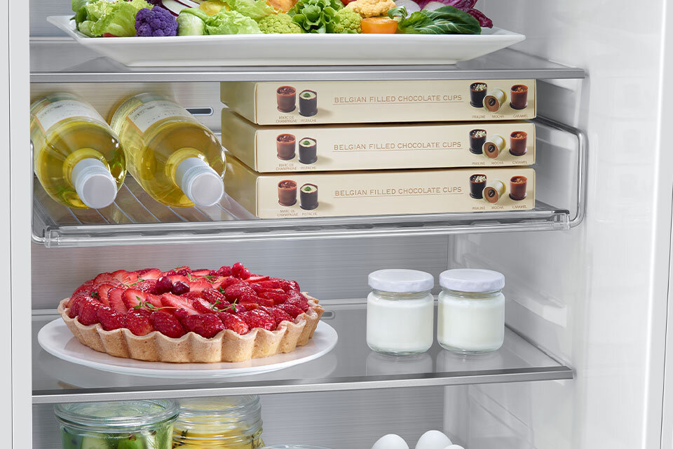 Wnętrze lodówki z widocznymi półkami, na których znajdują się różne produkty spożywcze, w tym butelki z napojami, pudełka z napisem \'BELGIAN FILLED CHOCOLATE CUPS\', tarta z truskawkami oraz słoiki z białą zawartością. Na górnej półce znajduje się talerz z warzywami.