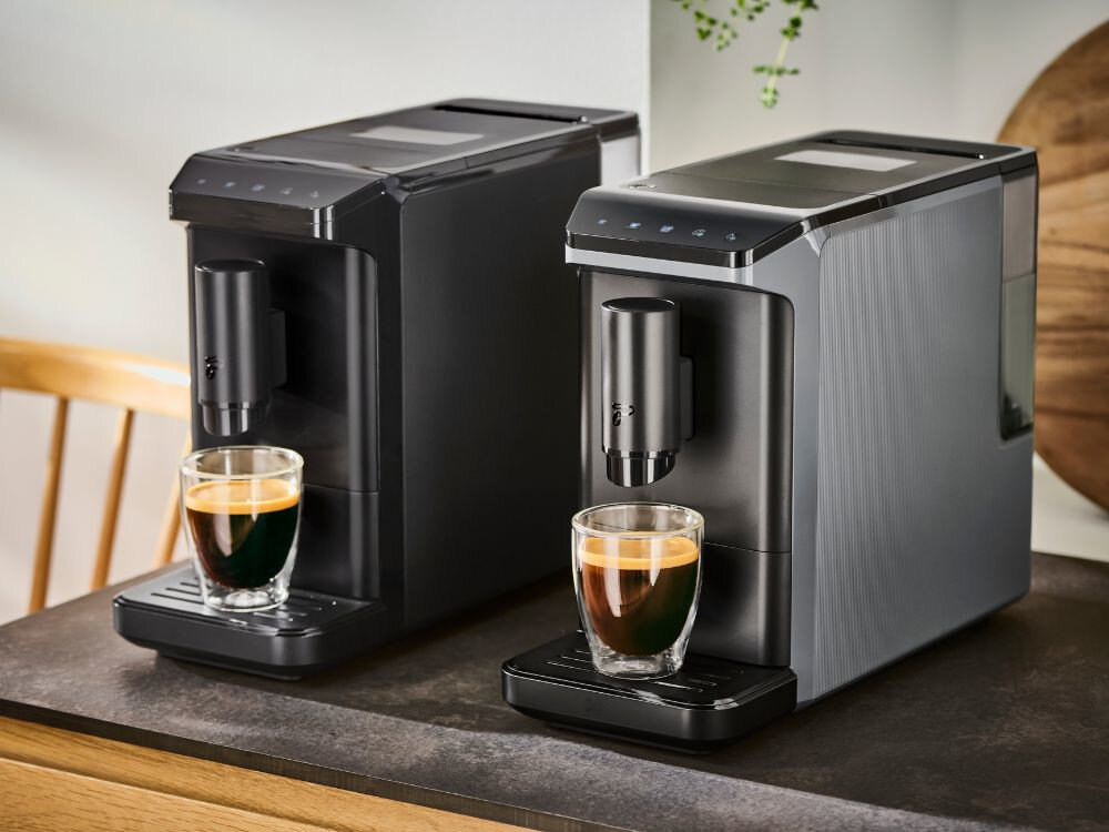 Na blacie stoją dwa czarne ekspresy do kawy, z których każdy przygotowuje filiżankę kawy. Ekspresy mają nowoczesny design i są ustawione obok siebie.