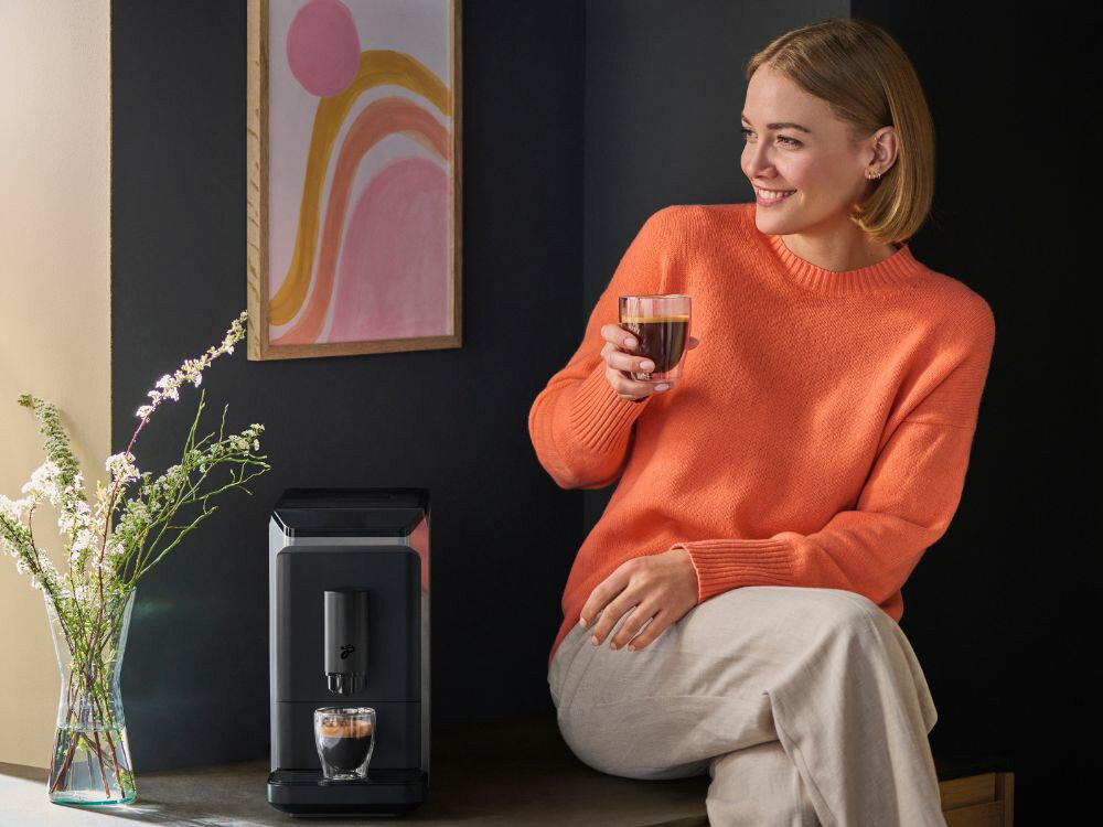 Na obrazie znajduje się czarny ekspres do kawy z filiżanką kawy na blacie. Obok siedzi osoba trzymająca szklankę z napojem, ubrana w pomarańczowy sweter.