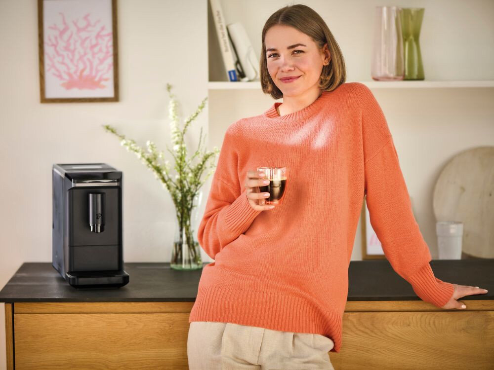 Na blacie stoi czarny ekspres do kawy. Obok znajduje się osoba trzymająca filiżankę z napojem.