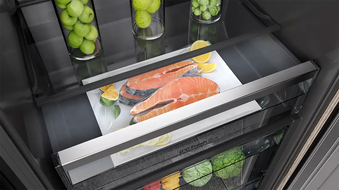 Wnętrze lodówki z szufladą zawierającą kawałki łososia na białej tacy, obok plasterków cytryny i limonki. W tle widoczne są szklane pojemniki z zielonymi jabłkami i brukselką oraz napis \'0°C Fresh Box\'.